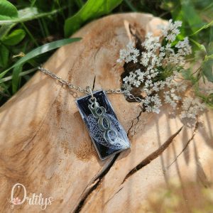 Un collier argenté, dont le médaillon rectangulaire a un fond noir, par dessus une papille argentée où il est écrit "boo" verticalement, et qui fait toute la hauteur du médaillon